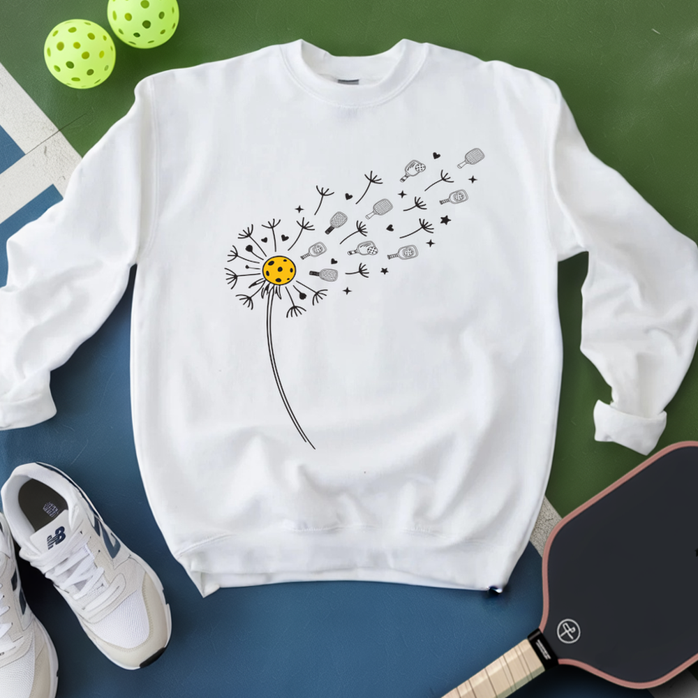 White sweatshirt with a dandelion graphic on a green and blue surface with pickleballs and shoes.