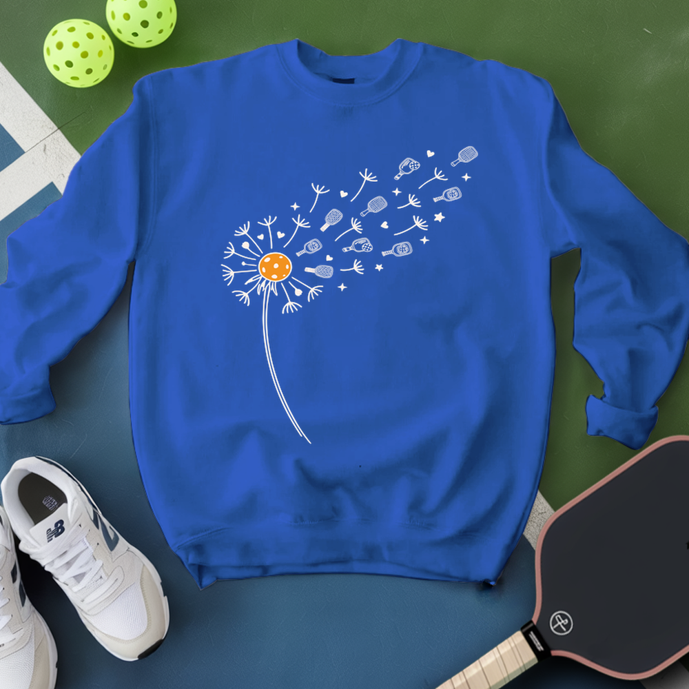 Blue sweatshirt with a dandelion graphic on a green surface with pickleballs and a paddle.