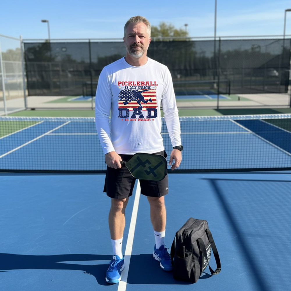 Man on a pickleball court wearing a white long-sleeve shirt with text and graphics, holding a paddle.