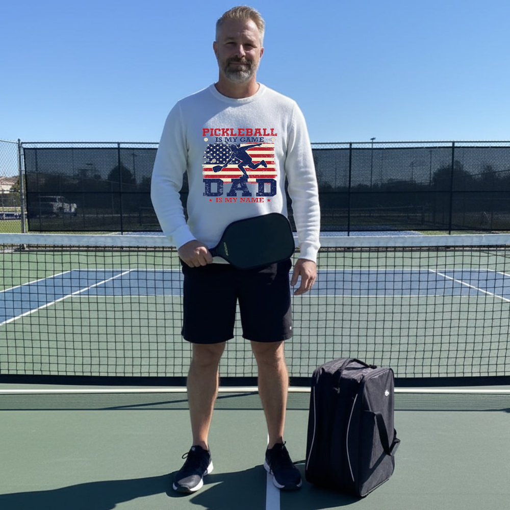 Man on a pickleball court wearing a sweatshirt with 'Pickleball Dad' text, holding a paddle.