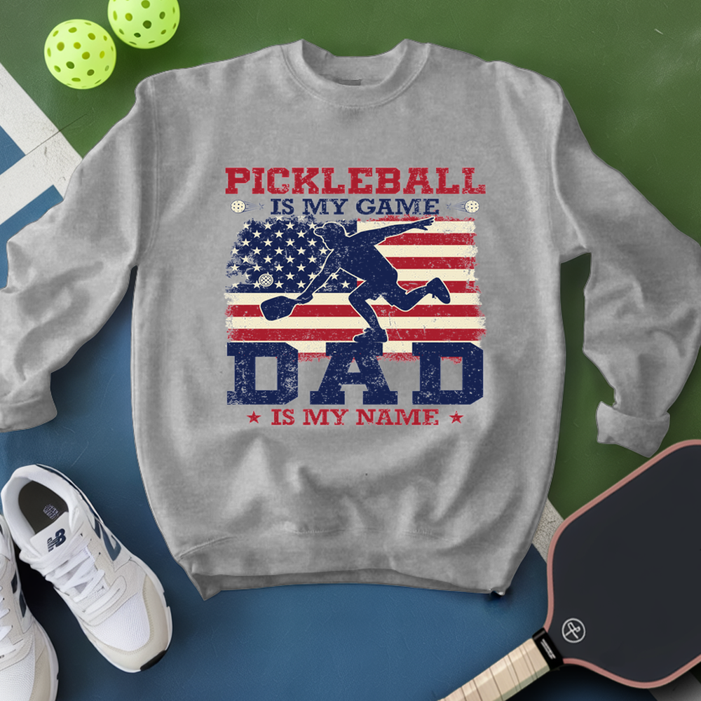 Gray sweatshirt with pickleball design and text, surrounded by pickleballs and a paddle on a green and blue background.