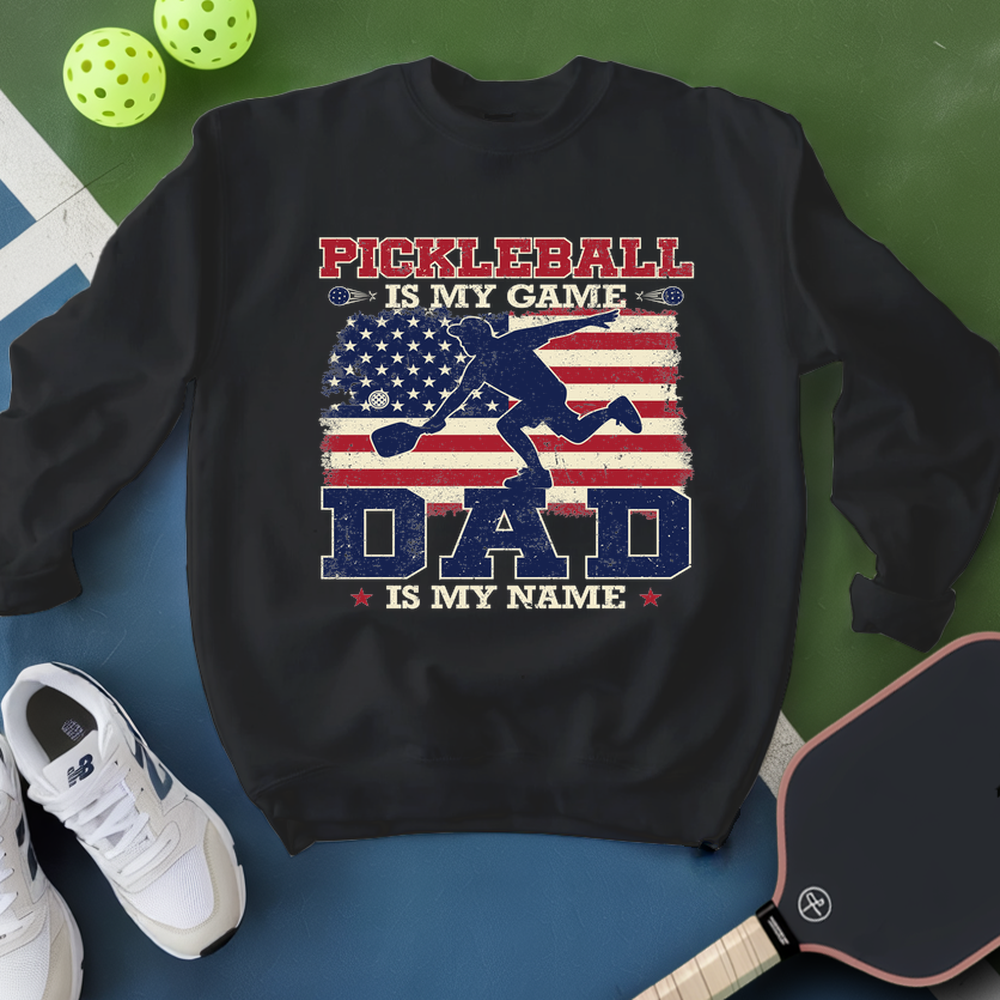 Black sweatshirt with pickleball design and text, surrounded by pickleballs and a paddle on a green and blue background.