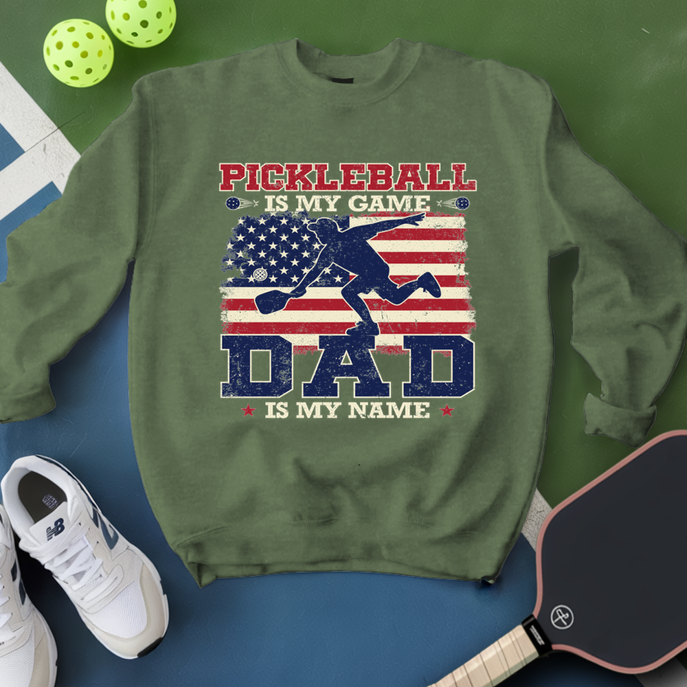 Green sweatshirt with pickleball design and text, surrounded by pickleballs and a paddle on a green and blue background.