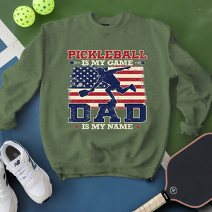 Green sweatshirt with pickleball design and text, surrounded by pickleballs and a paddle on a green and blue background.