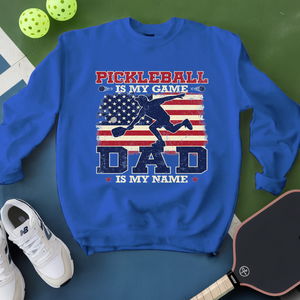 Blue sweatshirt with pickleball design and text on a green surface with pickleballs and shoes.