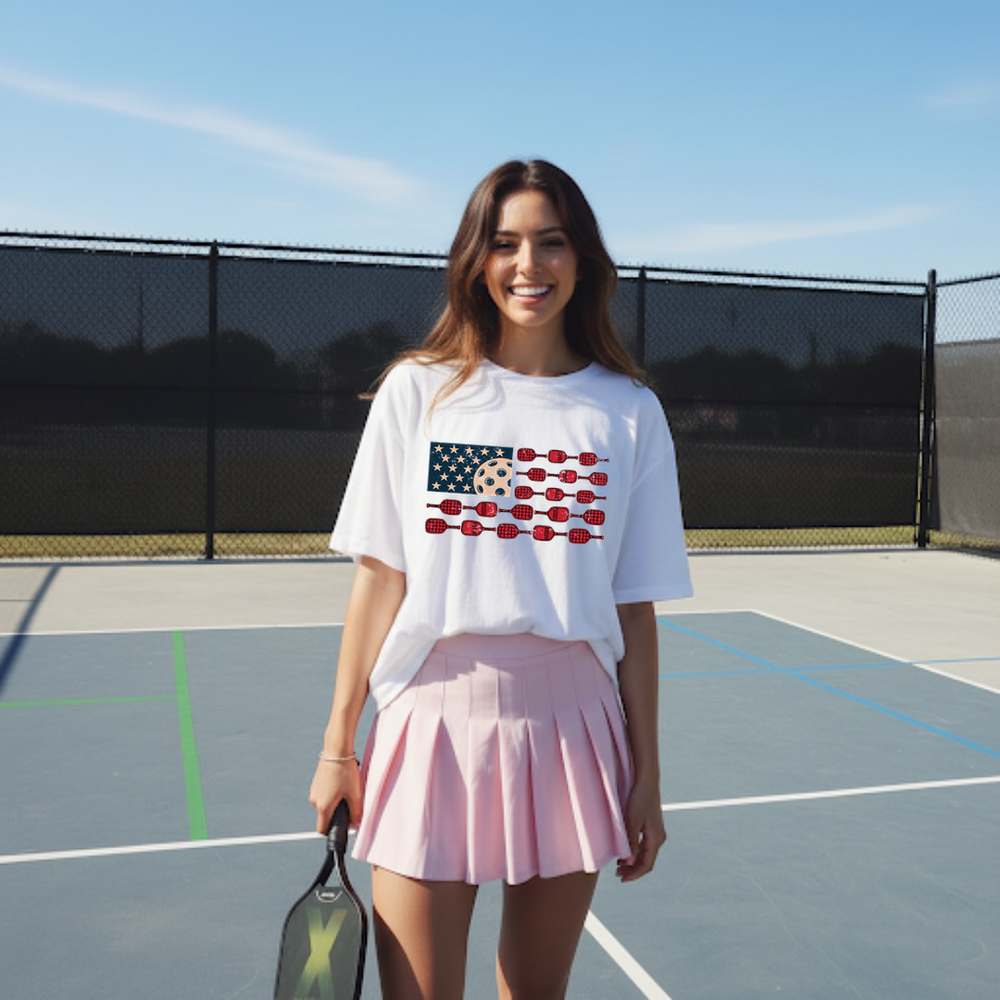 Woman on a tennis court wearing a white t-shirt with an American flag design and a pink skirt.