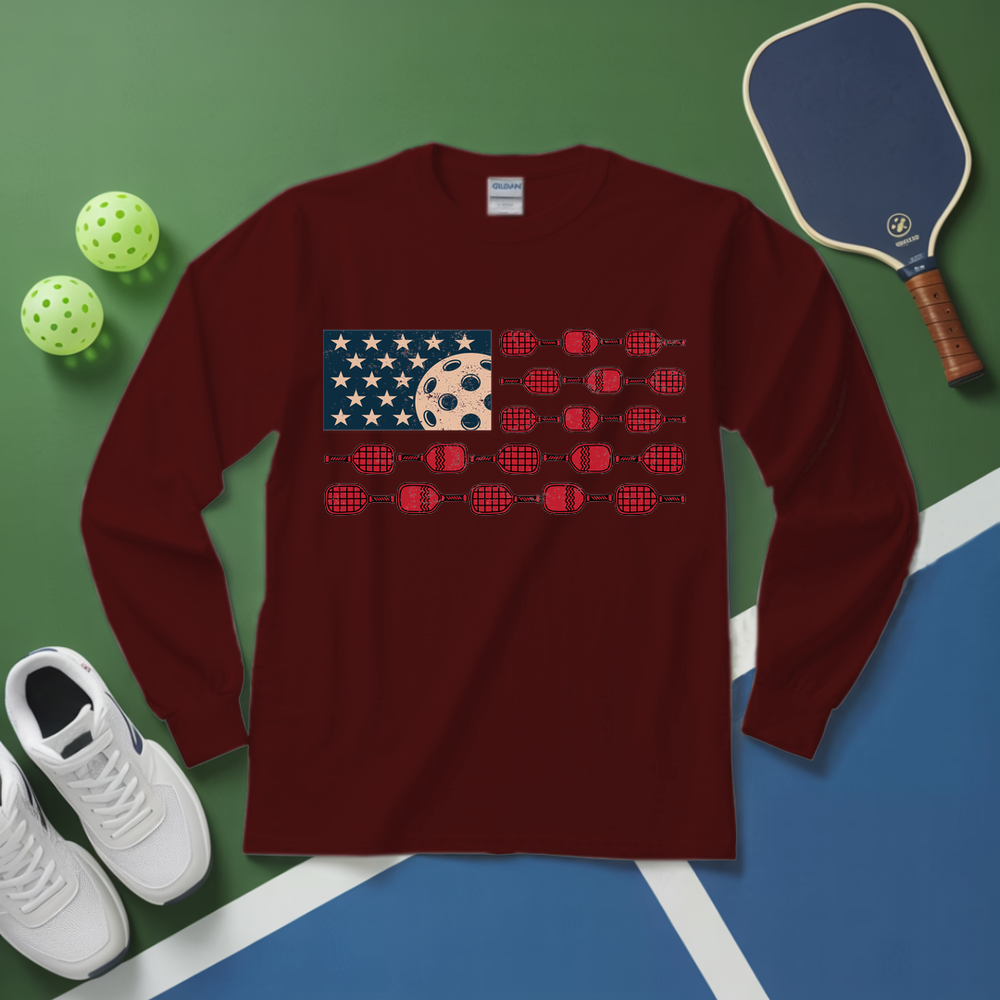 Maroon long-sleeve shirt with American flag and paddle graphic on a green and blue surface with pickleball equipment.