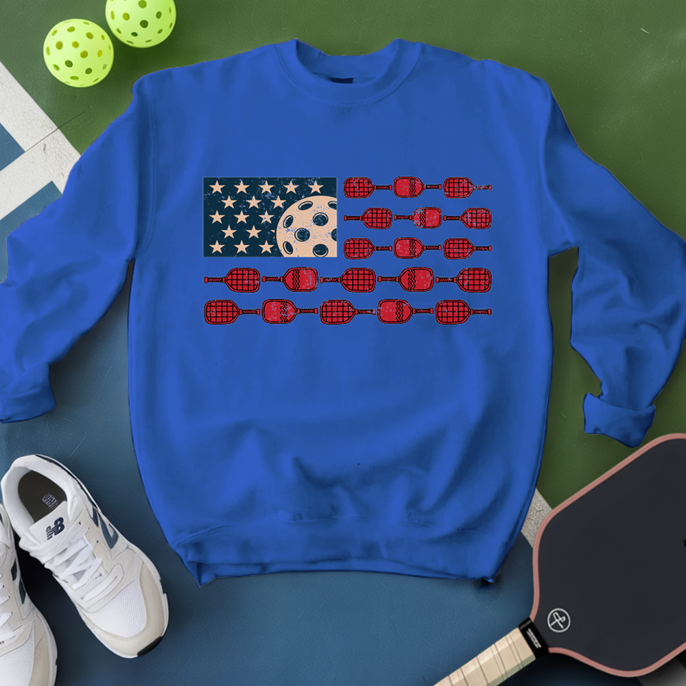 Blue sweatshirt with American flag design on a green surface with pickleball equipment.
