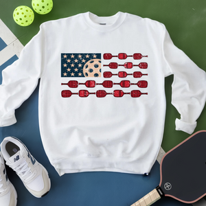 White sweatshirt with American flag design featuring pickleball elements on a green and blue surface with pickleballs and a paddle.