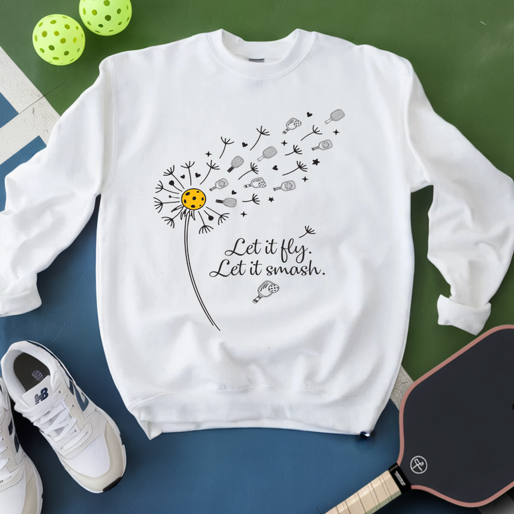 White sweatshirt with dandelion graphic and text, placed on a green and blue surface with pickleballs and a paddle.