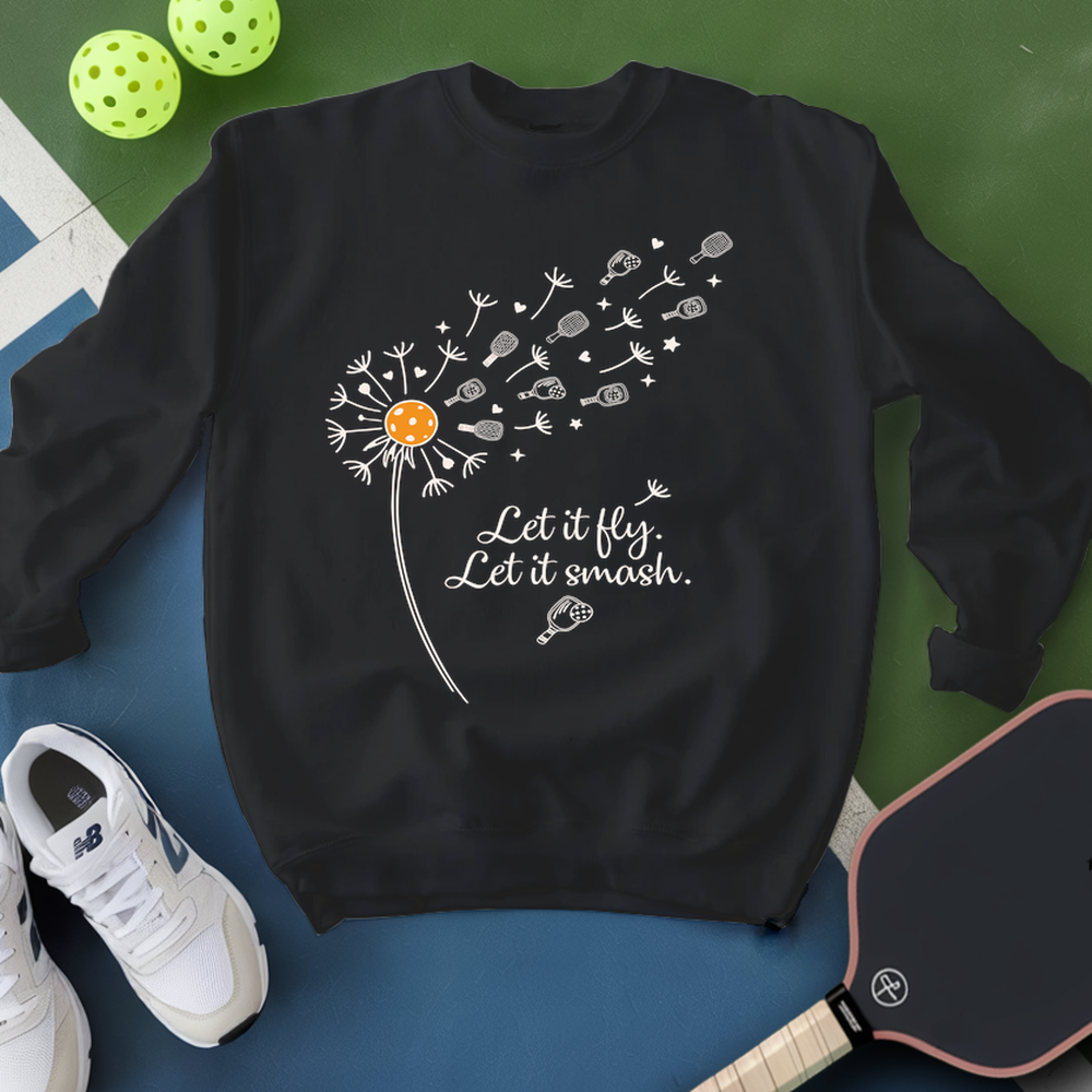 Black sweatshirt with a dandelion graphic and text, placed on a blue surface with pickleball equipment.
