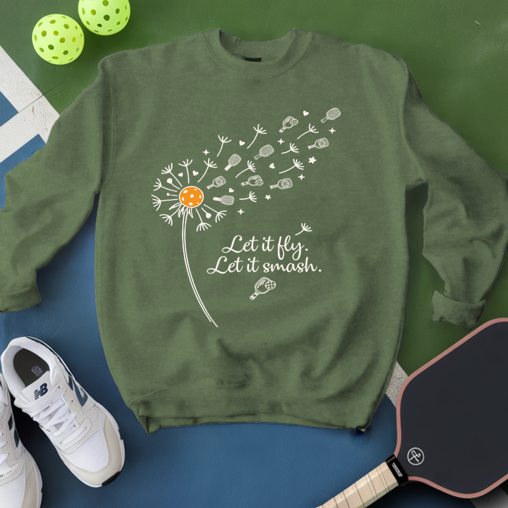 Green sweatshirt with dandelion graphic and text, placed on a blue surface with pickleball equipment.