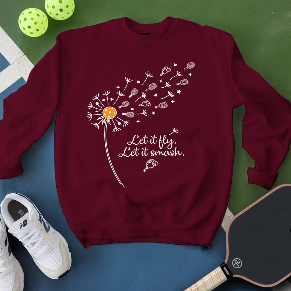 Maroon sweatshirt with dandelion graphic and text, placed on a blue and green surface with pickleball equipment.