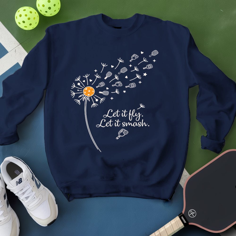 Navy blue sweatshirt with dandelion graphic and text, placed on a green and blue surface with pickleball equipment.