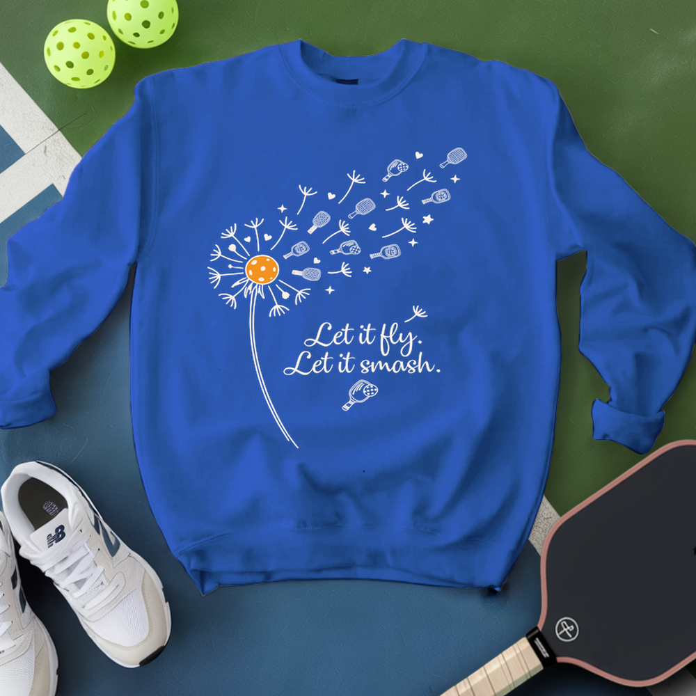 Blue sweatshirt with dandelion graphic and text, placed on a green surface with pickleball equipment.