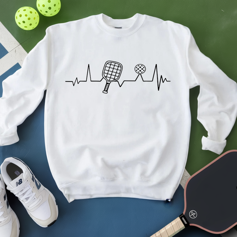 White sweatshirt with pickleball design on a green and blue surface with pickleballs and a paddle.
