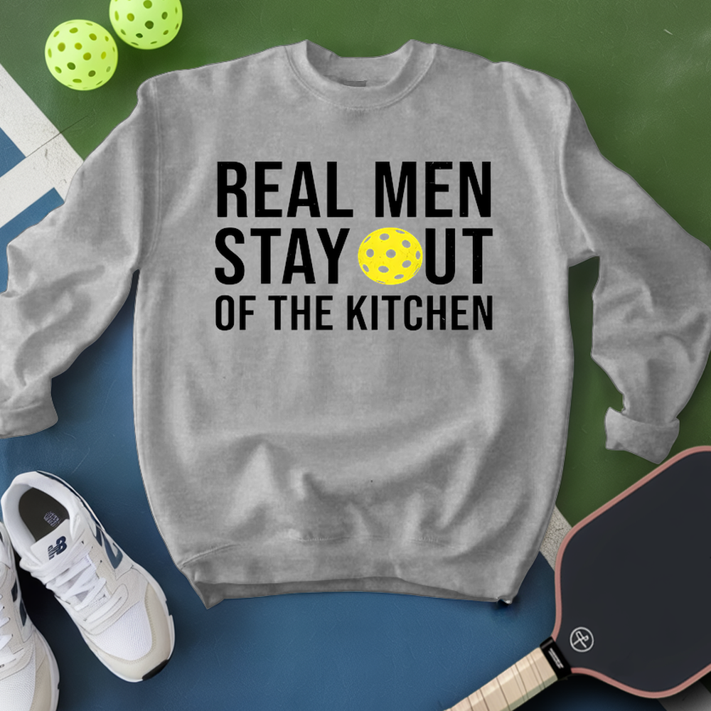 Gray sweatshirt with 'Real Men Stay Out of the Kitchen' text and pickleball ball, surrounded by pickleballs and a paddle on a green and blue background.