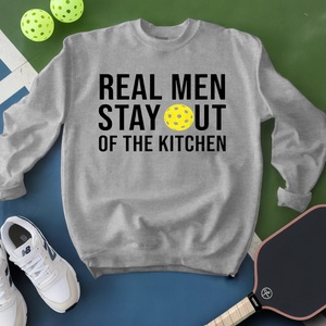 Gray sweatshirt with 'Real Men Stay Out of the Kitchen' text and pickleball ball, surrounded by pickleballs and a paddle on a green and blue background.