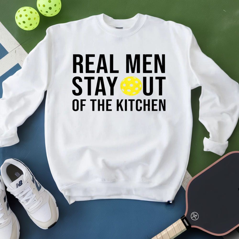 White sweatshirt with text 'Real men stay out of the kitchen' on a green and blue background with pickleball equipment.