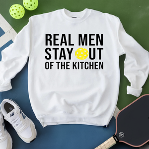 White sweatshirt with text 'Real men stay out of the kitchen' on a green and blue background with pickleball equipment.