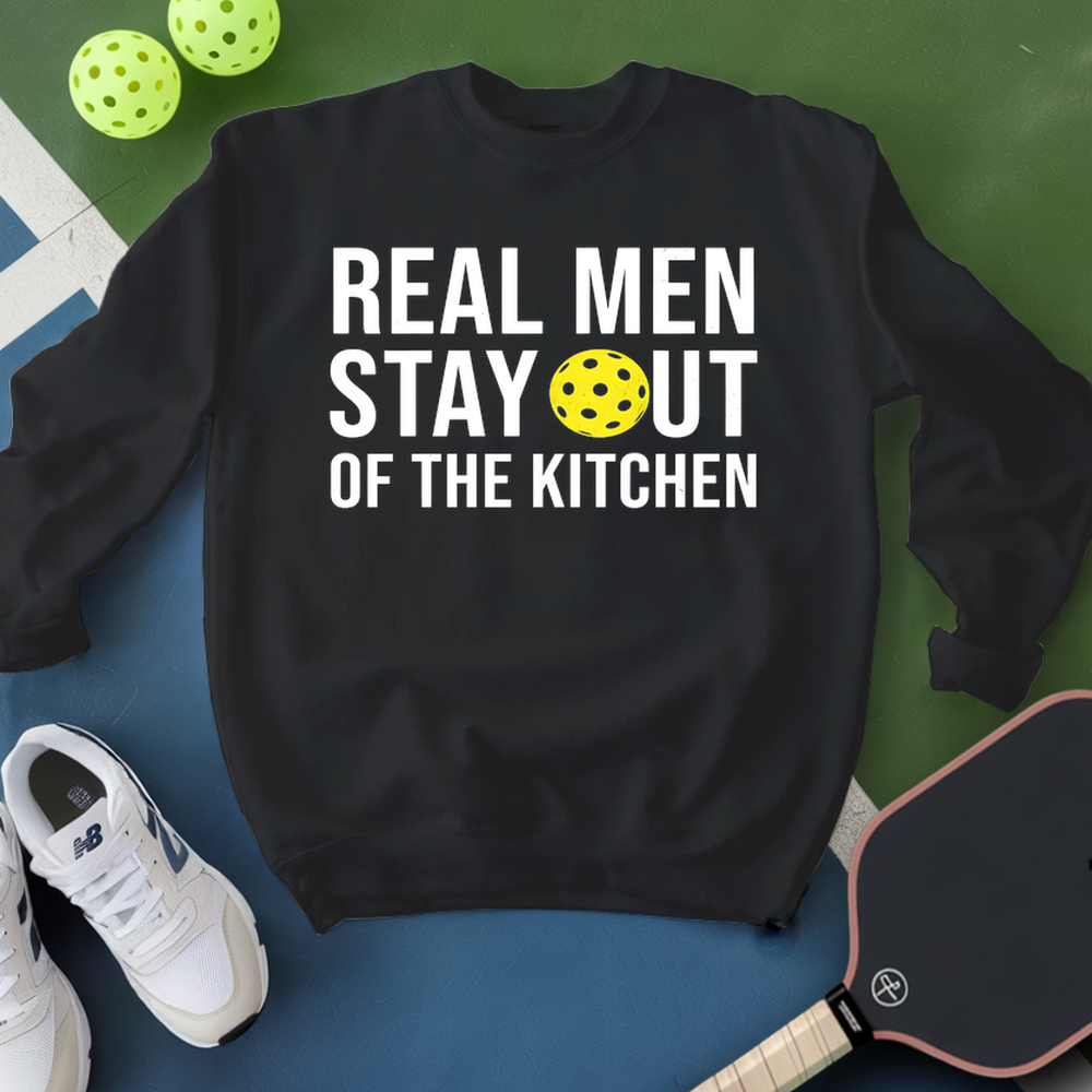 Black sweatshirt with 'Real Men Stay Out of the Kitchen' text and pickleball ball, surrounded by sports equipment on a green and blue background.