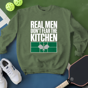 Green sweatshirt with 'Real Men Don't Fear The Kitchen' text, pickleball balls, paddle, and shoes on a green and blue background.