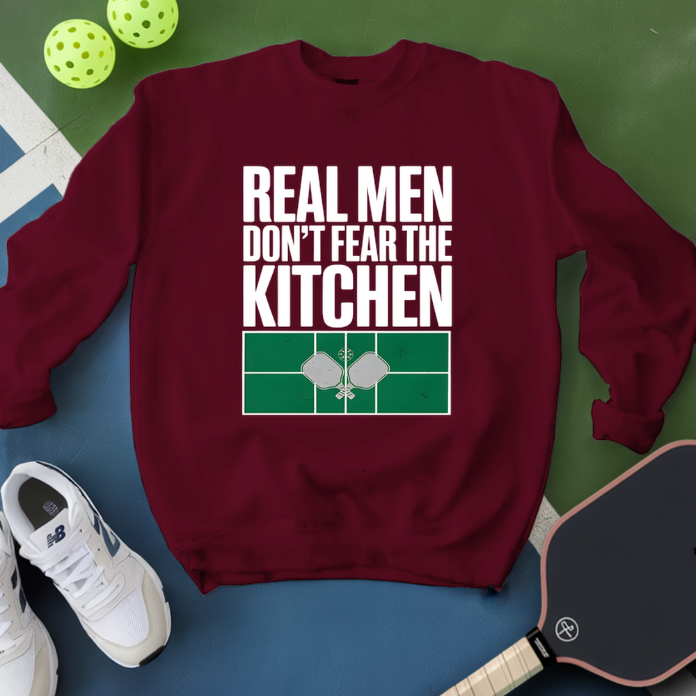 Maroon sweatshirt with 'Real Men Don't Fear The Kitchen' text on a green and blue background with pickleball equipment.