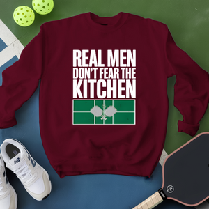 Maroon sweatshirt with 'Real Men Don't Fear The Kitchen' text on a green and blue background with pickleball equipment.