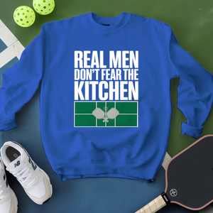 Blue sweatshirt with 'Real men don't fear the kitchen' text on a green surface with pickleball equipment.