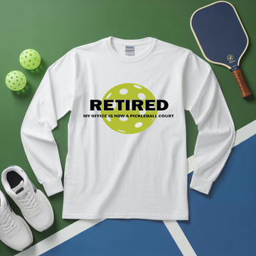 White long-sleeve shirt with pickleball-themed design on a green and blue surface with pickleball equipment.