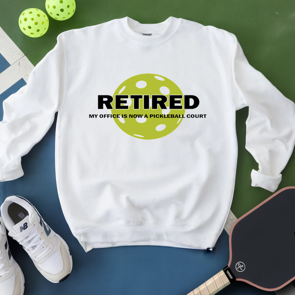 White sweatshirt with 'Retired' and pickleball ball design, next to pickleballs and a paddle on a green and blue background.