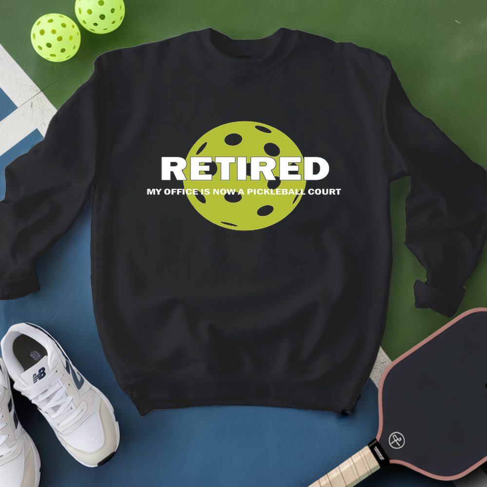 Black sweatshirt with pickleball design and text, surrounded by pickleballs and a paddle on a green and blue background.