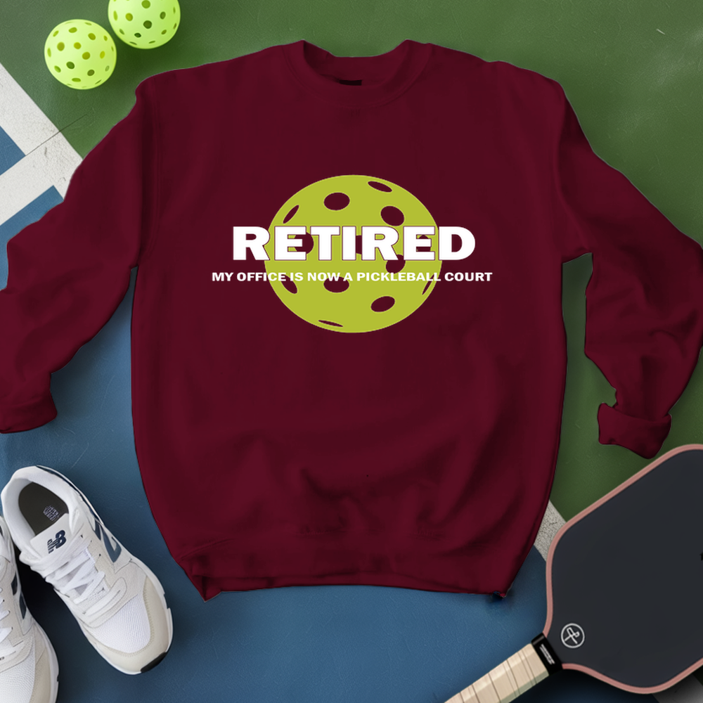 Maroon sweatshirt with pickleball design and text, surrounded by pickleballs and a paddle on a green and blue background.