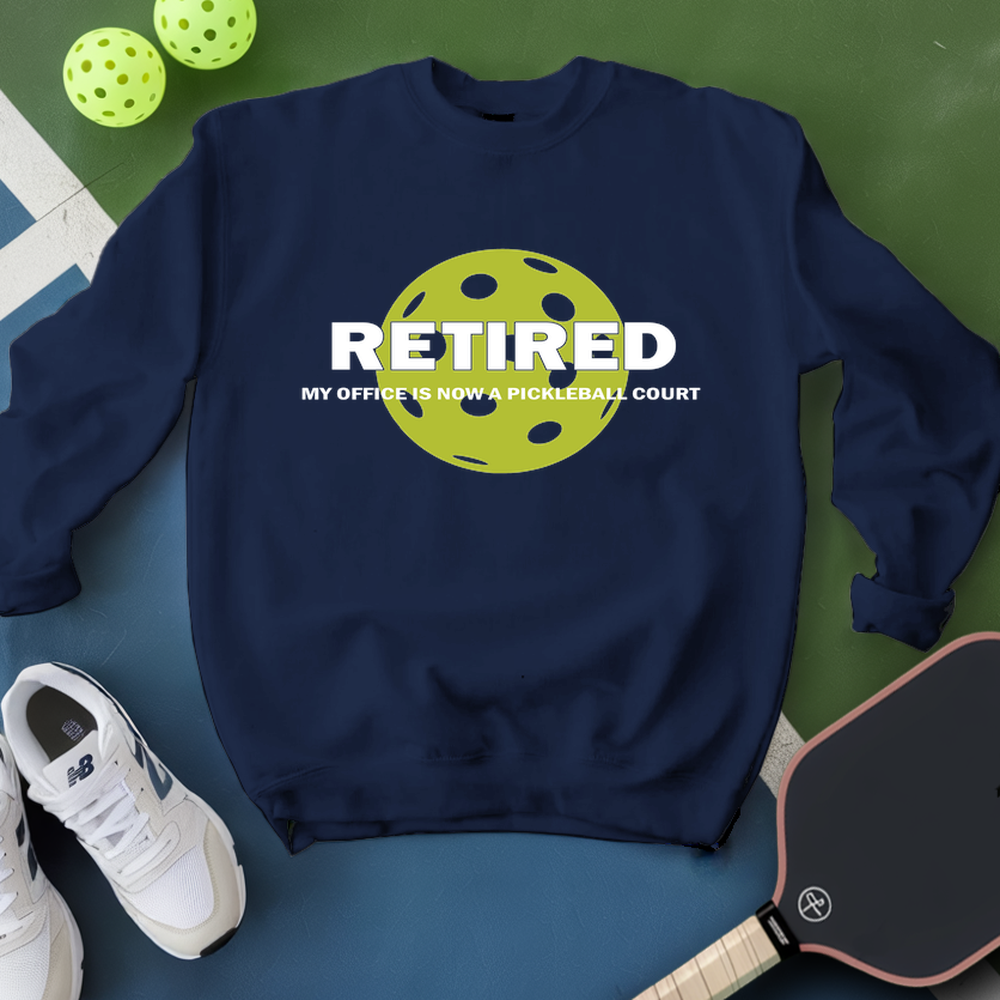 Navy sweatshirt with pickleball design and text, surrounded by pickleballs and a paddle on a green and blue background.