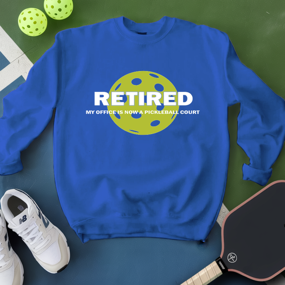 Blue sweatshirt with pickleball design and text on a green surface with pickleballs and a paddle.