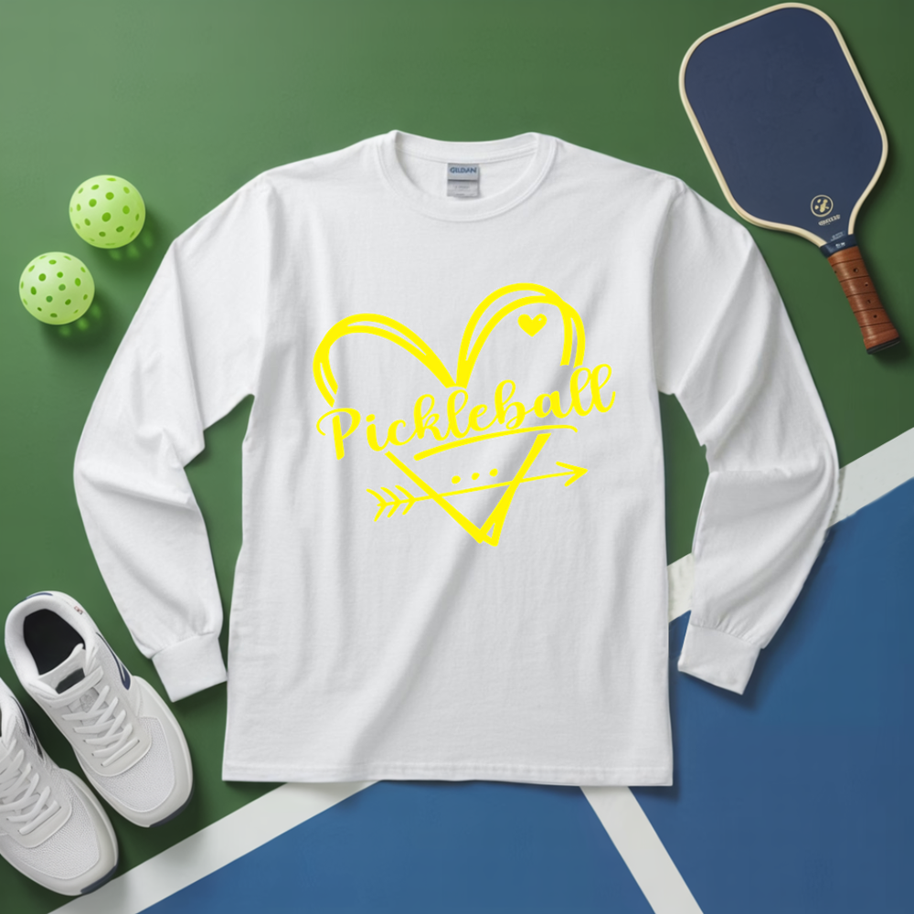 White long-sleeve shirt with yellow 'Pickleball' design on a green and blue surface with pickleball equipment.