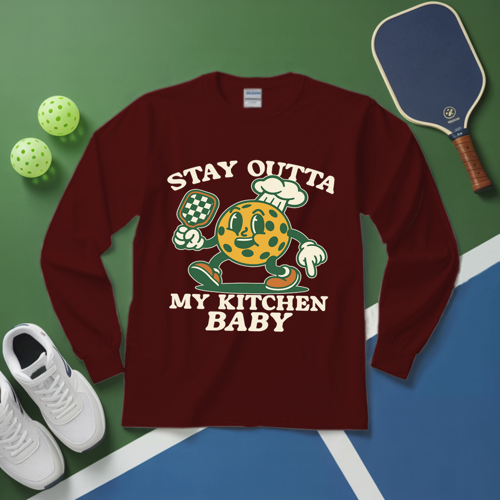 Maroon long-sleeve shirt with cartoon character and text on a green and blue surface with pickleball equipment.