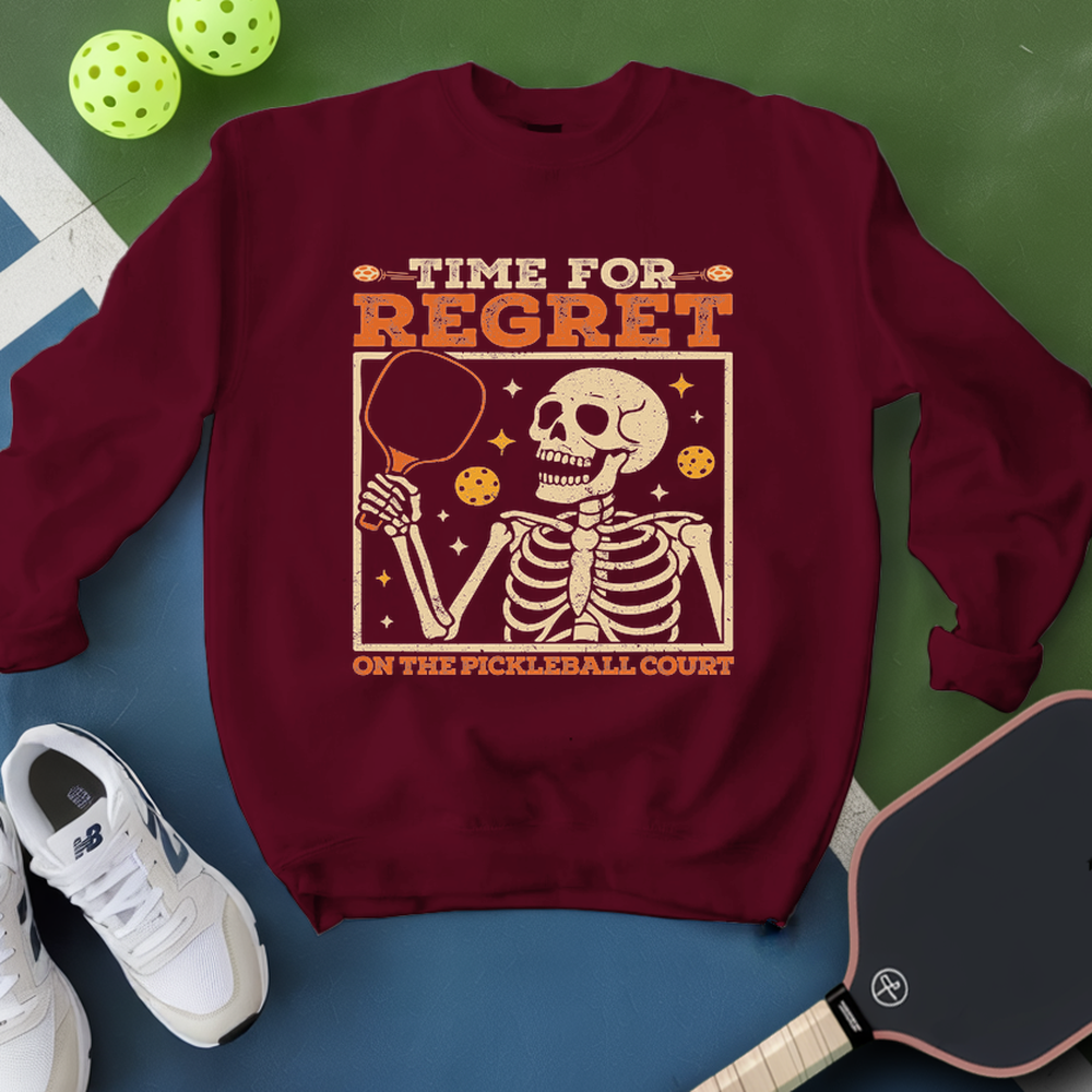 Maroon sweatshirt with skeleton graphic and pickleball text, placed on a blue and green surface with pickleballs and a paddle.