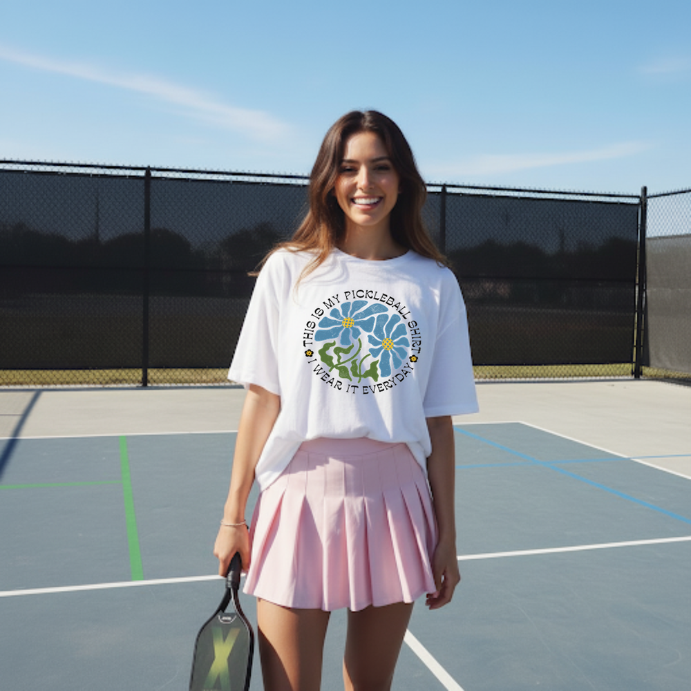 Woman on a tennis court wearing a white t-shirt with a graphic design and a pink skirt.