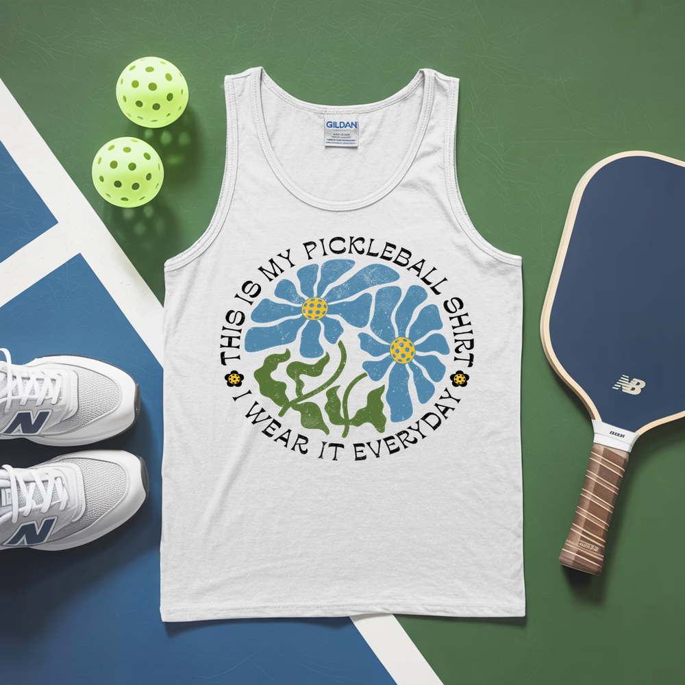 White tank top with pickleball-themed design, sneakers, and paddle on a green and blue surface.