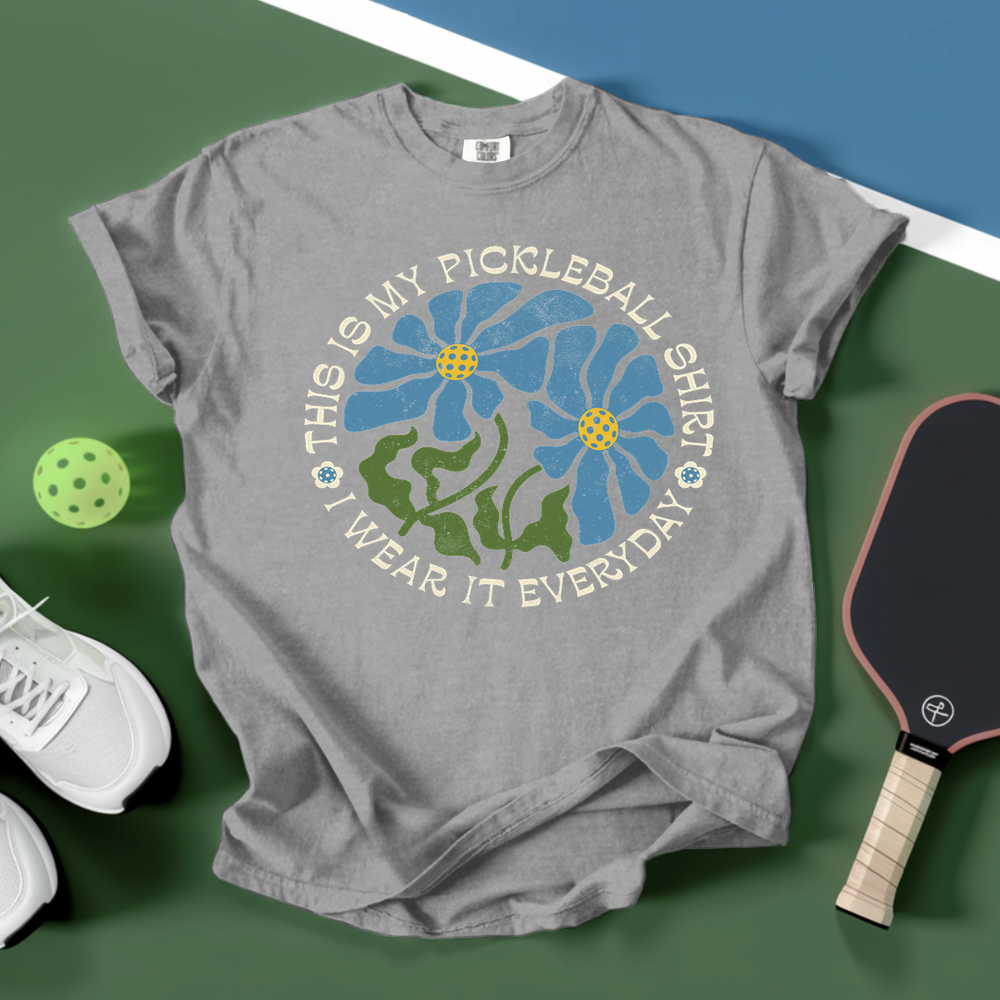 Gray t-shirt with pickleball-themed design on a green surface with paddle and ball.