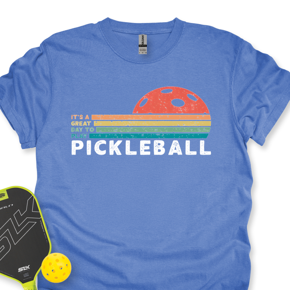 Blue t-shirt with pickleball graphic and text, next to a paddle and ball on a white background
