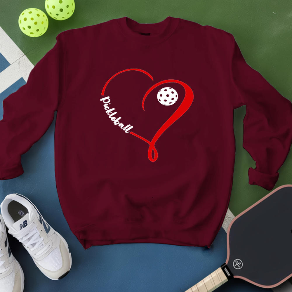 Maroon sweatshirt with red heart and pickleball design, next to a paddle and balls on a green and blue background.