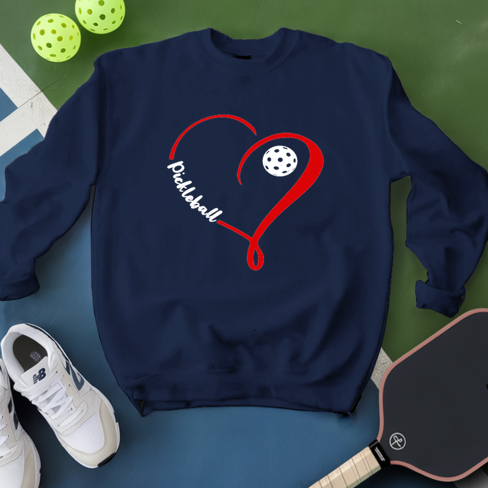 Navy blue sweatshirt with red heart and white pickleball design on a green and blue surface with pickleballs and a paddle.
