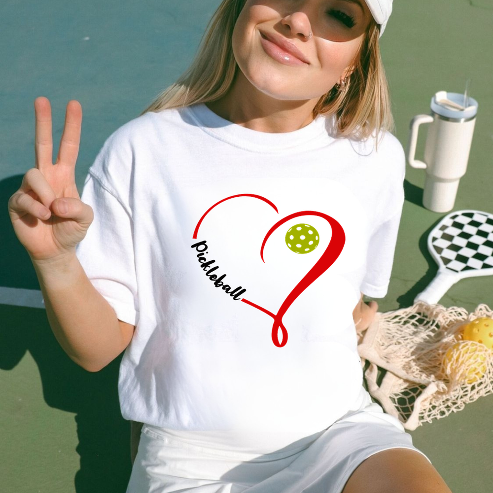 Woman wearing a white t-shirt with a pickleball design, making a peace sign on a tennis court.