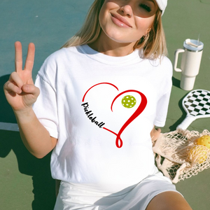 Woman wearing a white t-shirt with a pickleball design, making a peace sign on a tennis court.