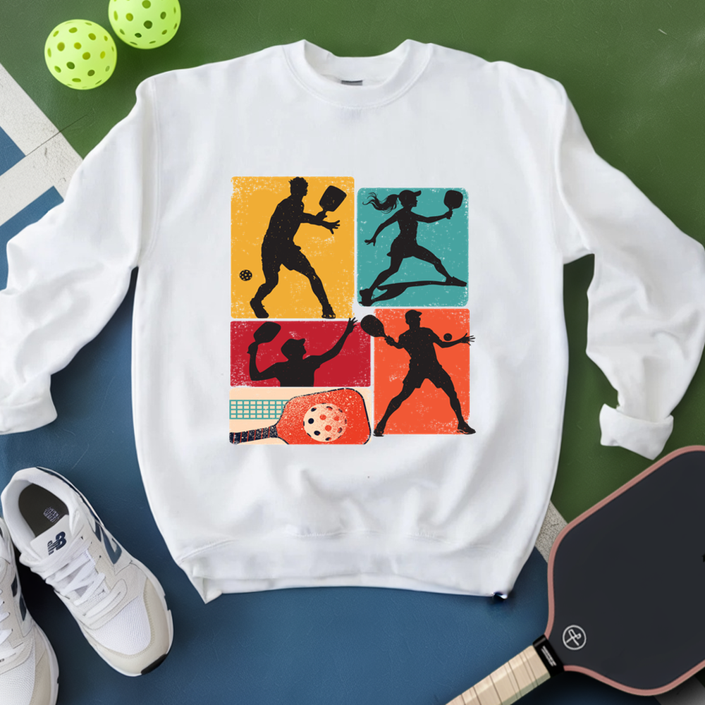 White sweatshirt with pickleball player design on a green and blue surface with pickleballs and a paddle.