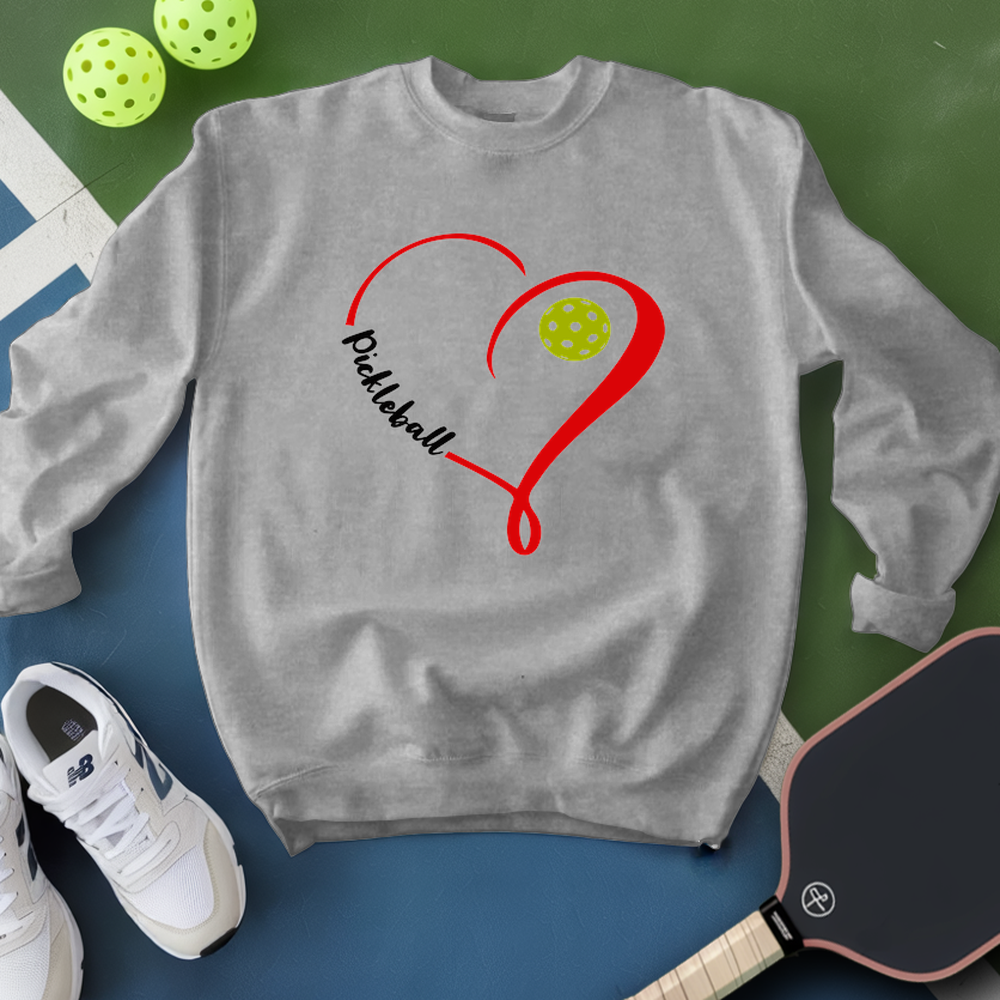 Gray sweatshirt with red heart and pickleball ball design, surrounded by pickleballs and a paddle on a green and blue background.