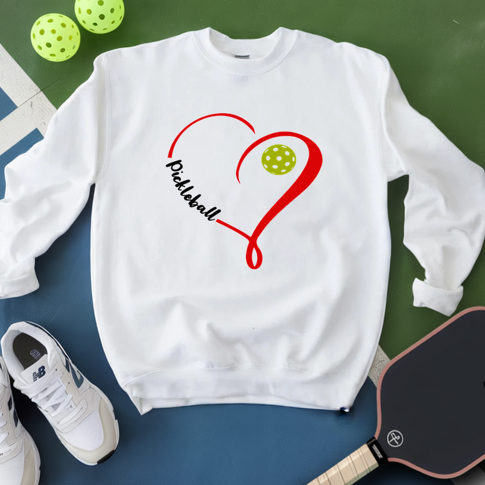 White sweatshirt with pickleball design on a green and blue surface with pickleballs and a paddle.