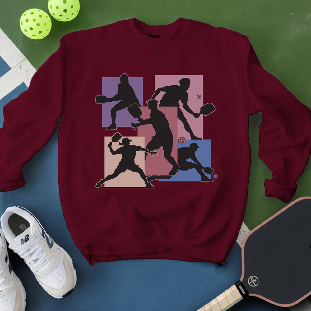 White sweatshirt with pickleball player design on a green and blue surface with pickleballs and a paddle.